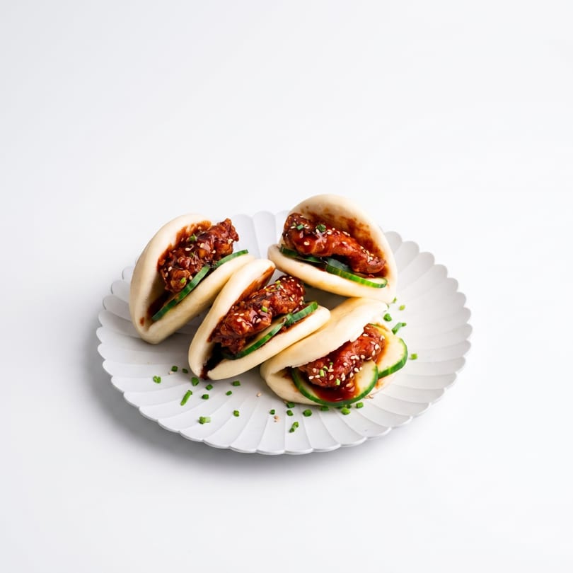 Four steamed bao buns with crispy chicken, cucumber, and sauce, garnished with chives on a white plate.